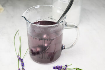 A glass measuring cup filled with purple liquid and a metal spoon is placed on a white surface, with lavender sprigs nearby.