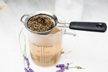A metal strainer with dried lavender sits atop a glass measuring cup containing light brown liquid, with lavender sprigs beside it on a white surface.