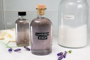 A glass bottle labeled Lavender Syrup sits on a counter next to a jar of sugar, a smaller bottle, and sprigs of fresh lavender.