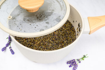 A cream-colored pot with a wooden handle and glass lid, partially open, containing dried lavender buds in water. Fresh lavender sprigs are placed beside the pot on a white surface.