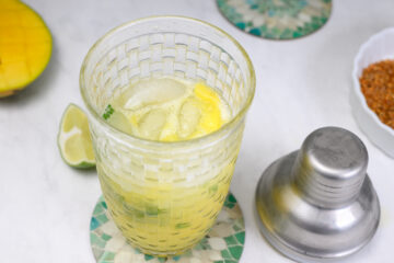 A glass with a yellow drink, ice, lime, and mango chunks sits on a coaster next to a metal cocktail shaker and a bowl of chili powder on a white surface.