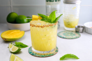 A yellow cocktail with ice in a glass rimmed with chili powder, garnished with mango, lime, and mint. Limes and mango are on the table, with a bottle and shaker in the background.