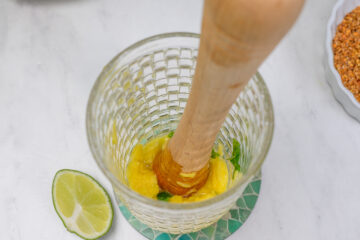 A wooden muddler presses pineapple and mint in a glass; a lime slice sits nearby on a green-patterned coaster, with a dish of seeds partially visible on the side.