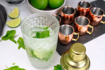 A glass of mint and lime cocktail with ice sits next to a gold shaker, copper mugs, lime wedges, and a bowl of whole limes on a white surface.
