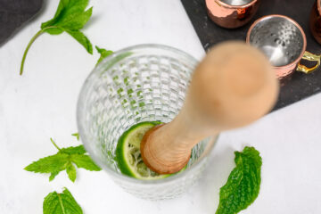 A muddler presses a lime slice in a textured glass, surrounded by fresh mint leaves on a white surface.
