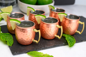 Six copper mugs with lime wedges and drinks inside are arranged on a black slate tray, surrounded by mint leaves.