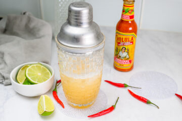 A cocktail shaker filled with a yellow drink sits on a coaster, surrounded by lime slices, red chili peppers, and a bottle of Cholula hot sauce on a white countertop.
