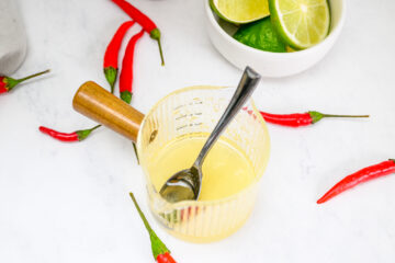 A glass measuring cup with a spoon and yellow liquid, surrounded by red chili peppers and a bowl of lime halves on a white surface.