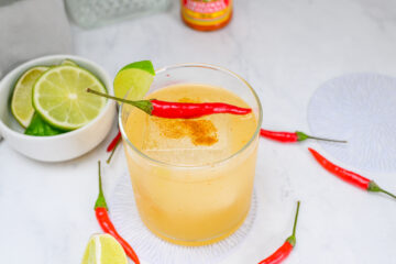 A glass of light yellow cocktail with ice, garnished with a red chili pepper and lime slice. Lime wedges and whole red chilies are arranged on a white surface nearby.