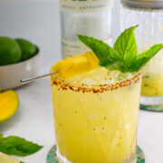 A yellow cocktail garnished with mint and mango chunks, rimmed with chili powder, sits on a coaster next to lime wedges, a shaker, mango, and a bottle in the background.