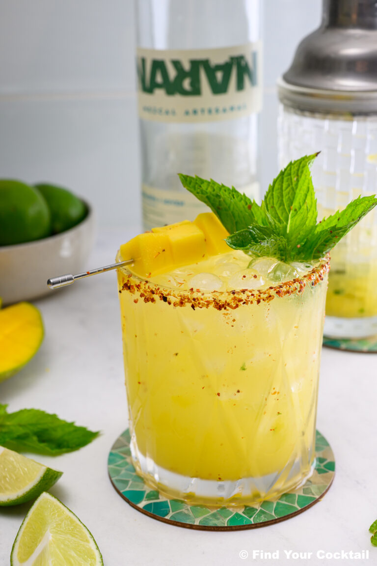 A yellow cocktail garnished with mint and mango chunks, rimmed with chili powder, sits on a coaster next to lime wedges, a shaker, mango, and a bottle in the background.