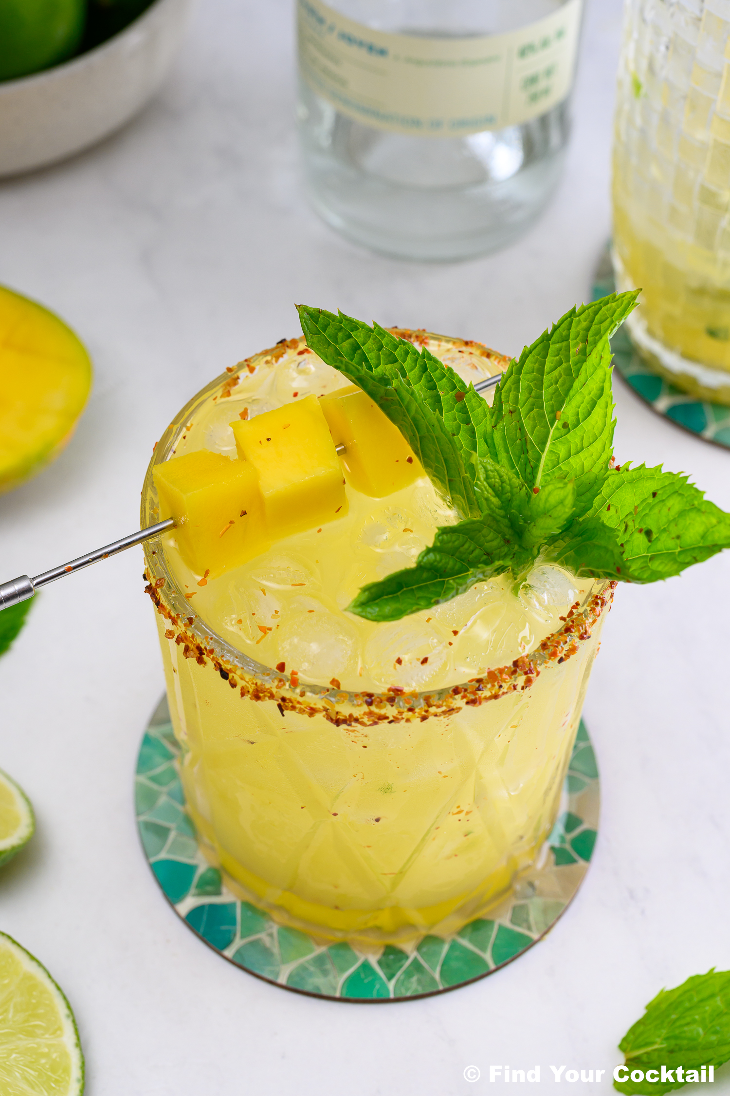 A yellow cocktail with ice, chili powder rim, mint sprig, and mango cubes on a pick, served on a mosaic coaster.