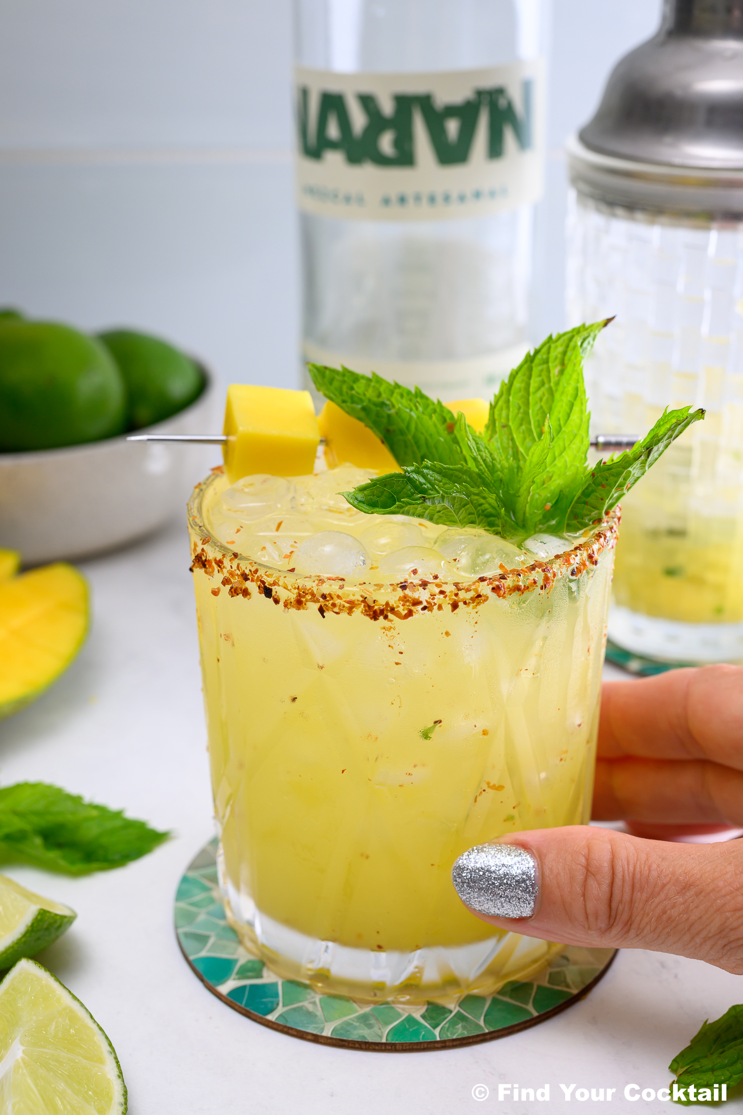 A hand holds a glass of yellow cocktail garnished with mint and mango cubes, with a chili-rimmed edge. Limes, mango, and a bottle are in the background.