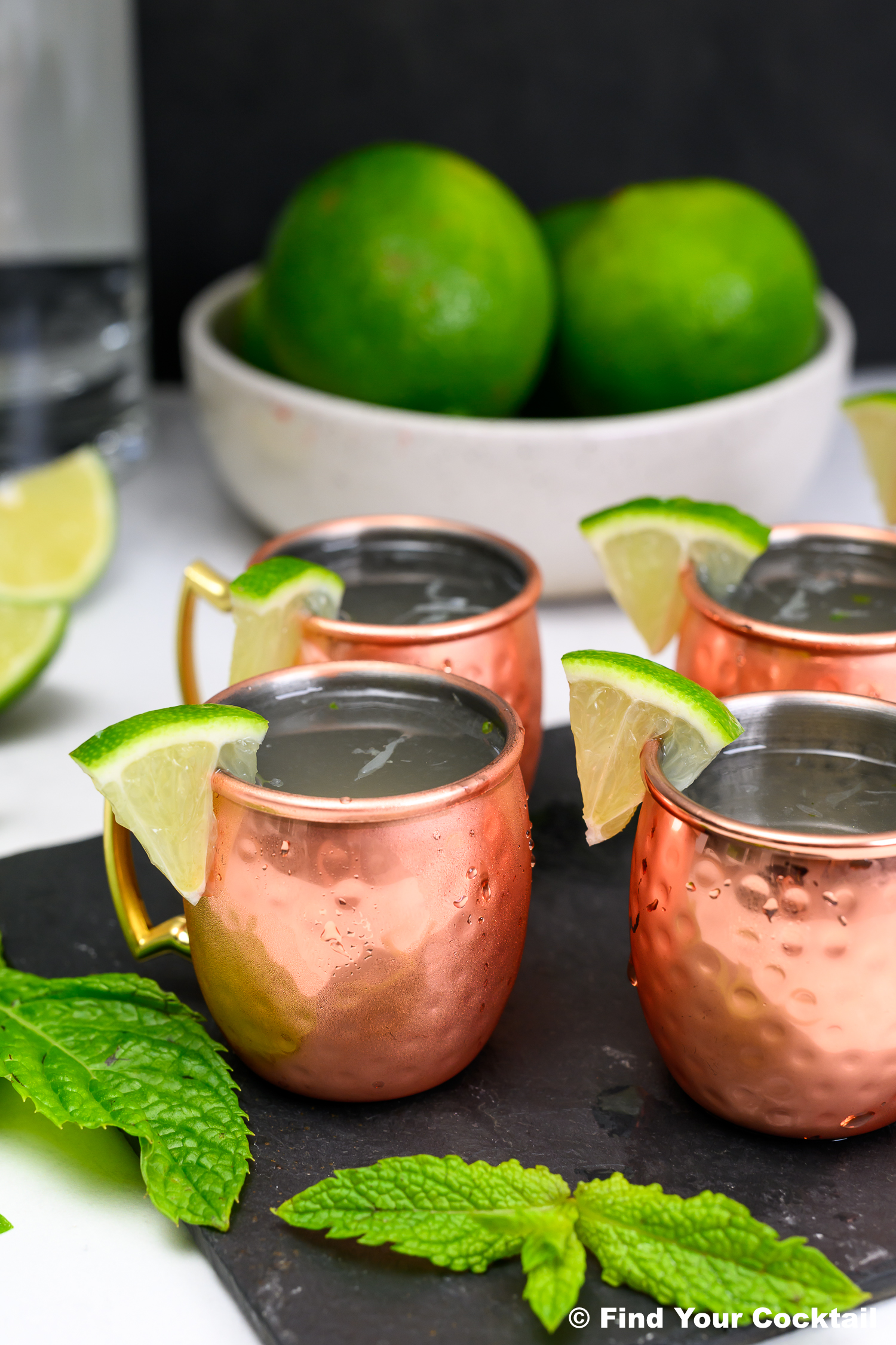 Four copper mugs filled with drinks, garnished with lime wedges, sit on a slate board with fresh mint and whole limes in the background.