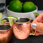 A hand with glittery nail polish holds a small copper mug with a lime wedge; more mugs, limes, and mint leaves are in the background.