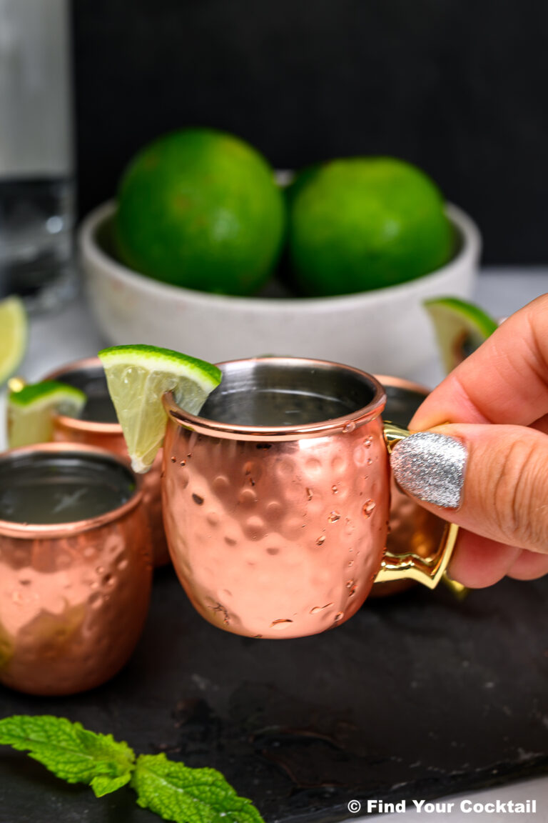 A hand with glittery nail polish holds a small copper mug with a lime wedge; more mugs, limes, and mint leaves are in the background.