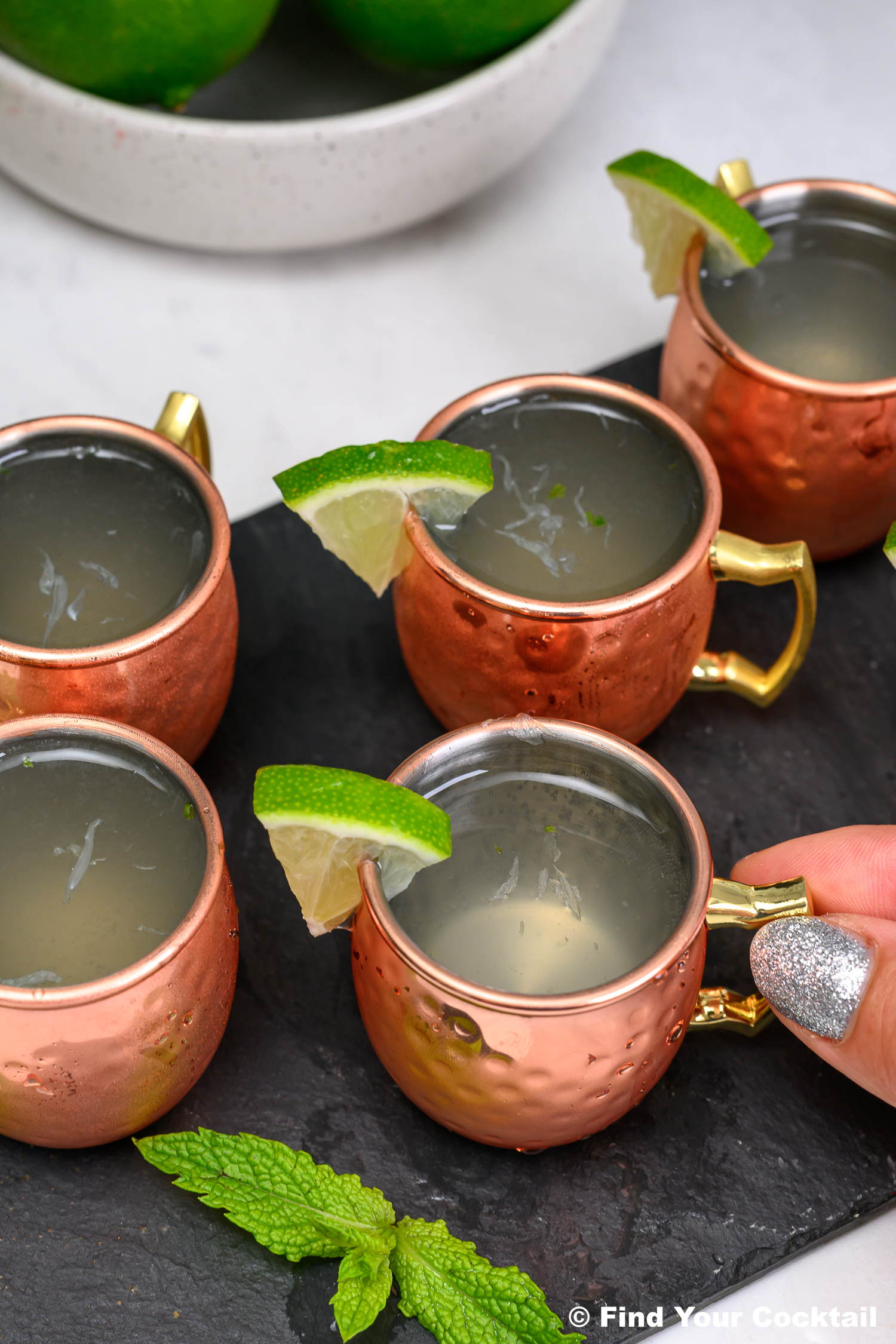 Five small copper mugs filled with cocktails, each garnished with a lime wedge, are arranged on a slate surface with mint leaves nearby. A hand touches one mug.