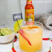 A cocktail garnished with a red chili pepper and lime wedge, surrounded by lime slices, whole red chilies, a bottle of Cholula hot sauce, and a bottle of 1800 tequila.