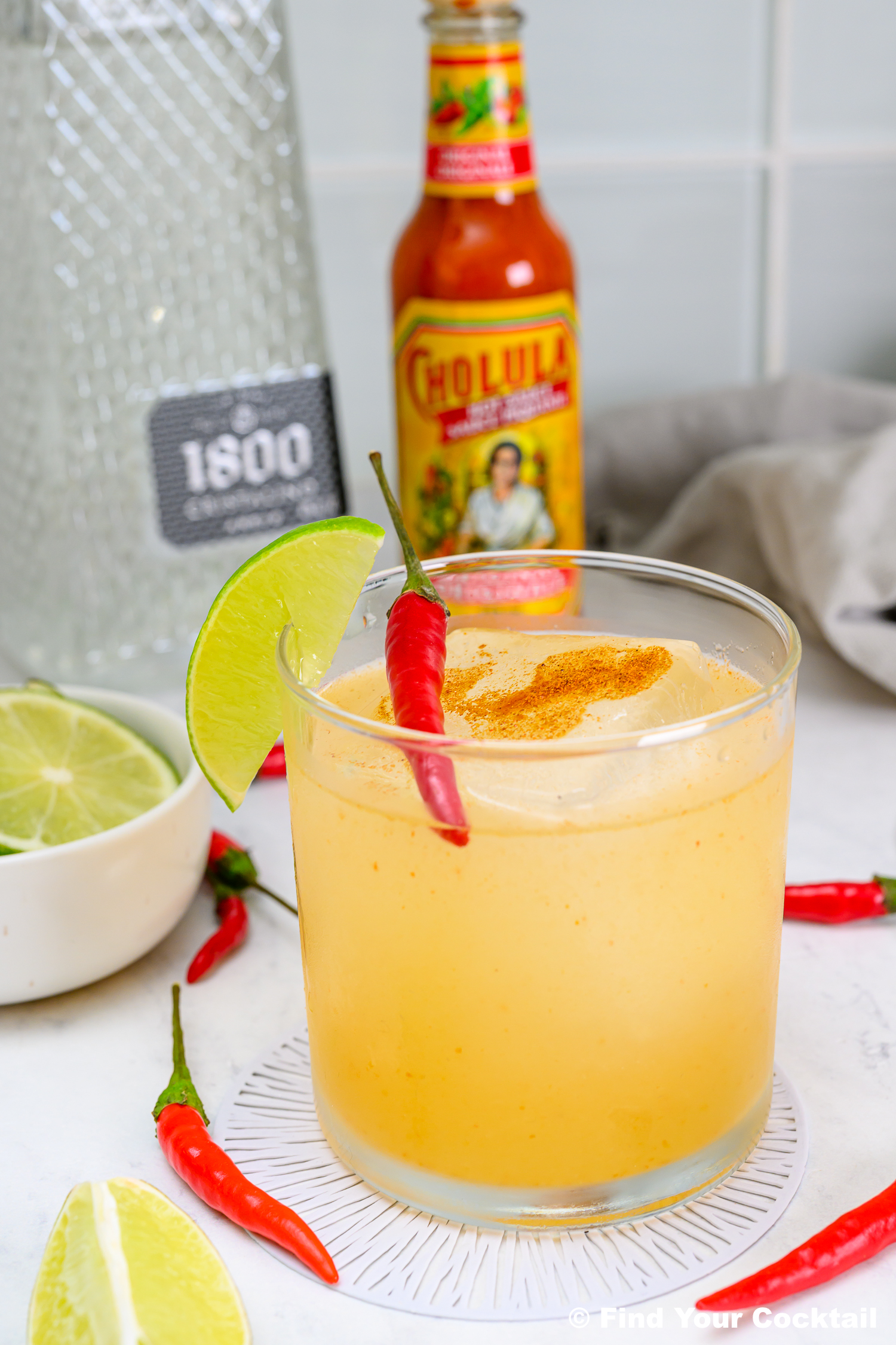 A cocktail garnished with a lime wedge, chili pepper, and spice sits on a coaster. Cholula hot sauce, tequila bottle, and sliced limes are in the background.