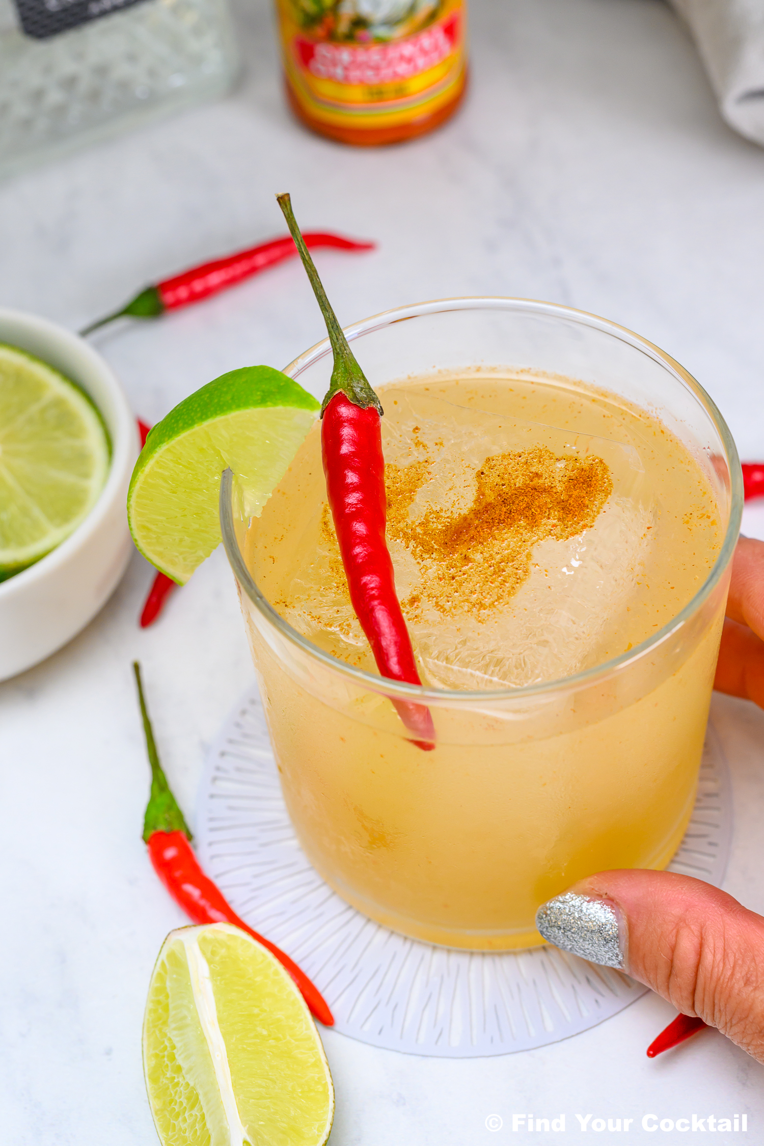 A hand holds a cocktail garnished with a lime wedge and red chili pepper, with a bowl of lime slices and loose chilies on a white surface.
