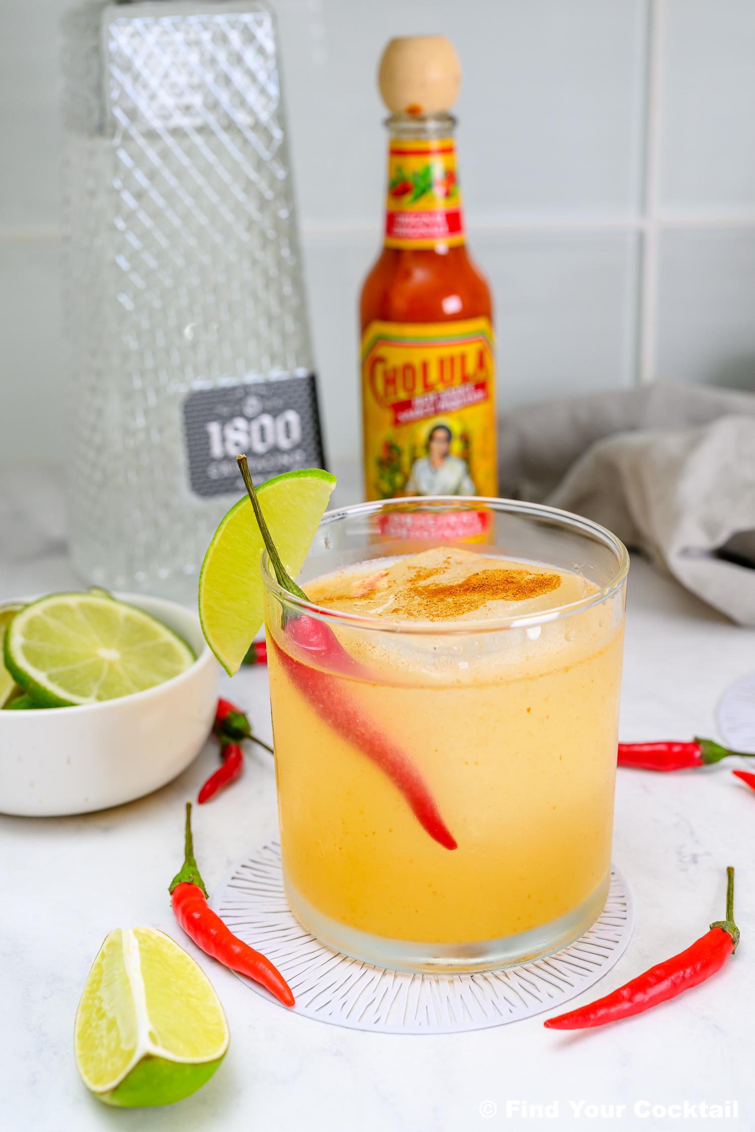 A cocktail garnished with a red chili pepper and lime wedge, surrounded by lime slices, whole red chilies, a bottle of Cholula hot sauce, and a bottle of 1800 tequila.