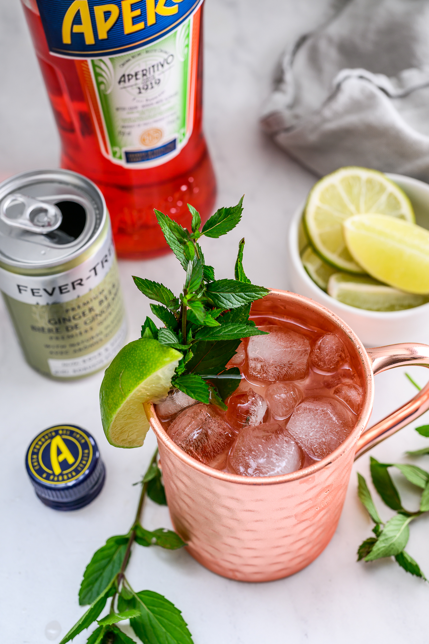 A copper mug filled with ice, garnished with mint and a lime wedge, sits next to a can of Fever-Tree, a bottle of Aperitivo, lime slices, and mint sprigs.