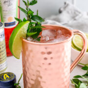 A copper mug filled with an iced cocktail, garnished with a lime wedge and mint sprig, sits on a white surface with Aperol, a bottle cap, and fresh mint nearby.