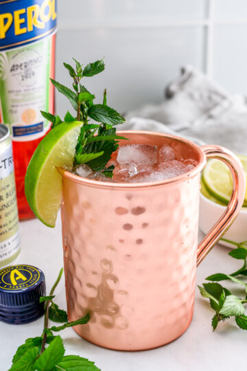 A copper mug filled with an iced cocktail, garnished with a lime wedge and mint sprig, sits on a white surface with Aperol, a bottle cap, and fresh mint nearby.