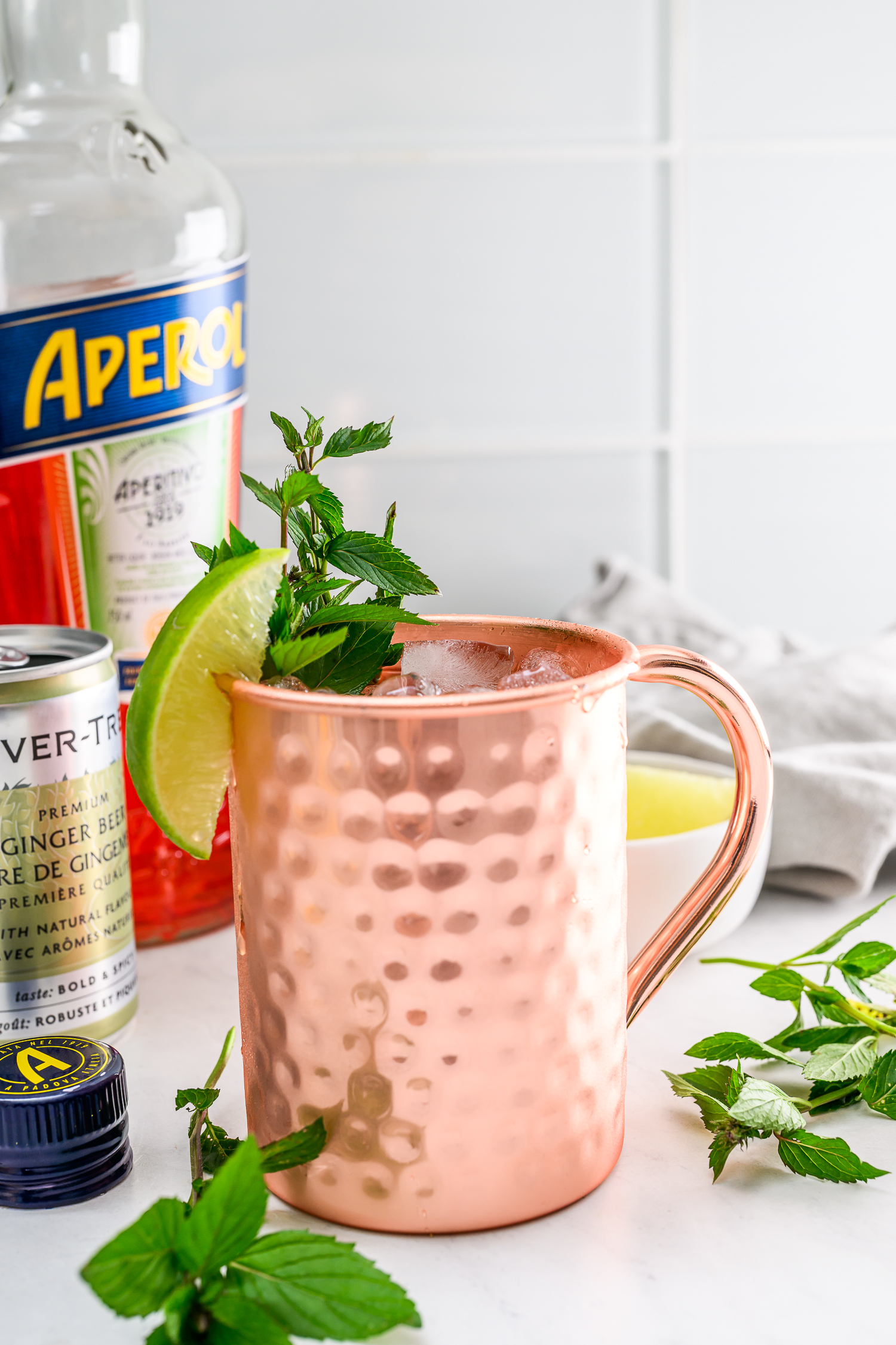 A copper mug filled with a cocktail, garnished with mint and a lime wedge, sits next to a bottle of Aperol, a can of ginger beer, and fresh mint on a white surface.