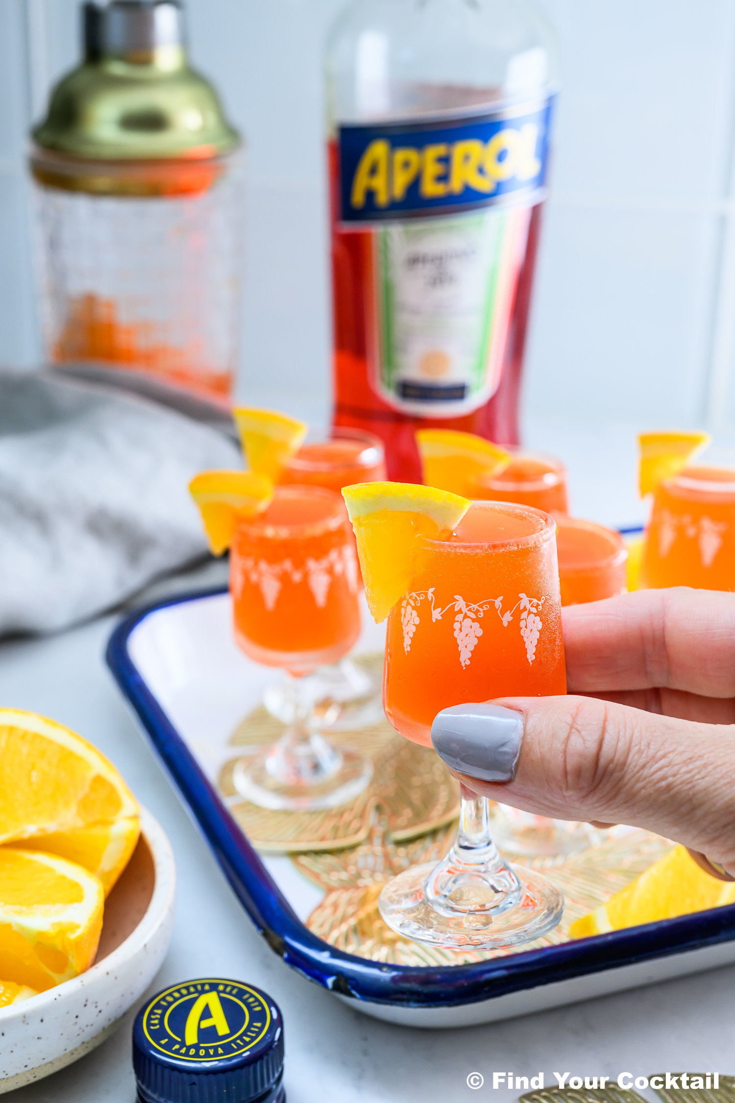 A hand holds a small glass of orange Aperol cocktail garnished with an orange slice; other filled glasses, a bottle of Aperol, and orange slices are visible in the background.