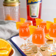 A tray of orange Aperol cocktails in small glasses with orange slices, a shaker, a bottle of Aperol, and a bowl of orange wedges in the background.