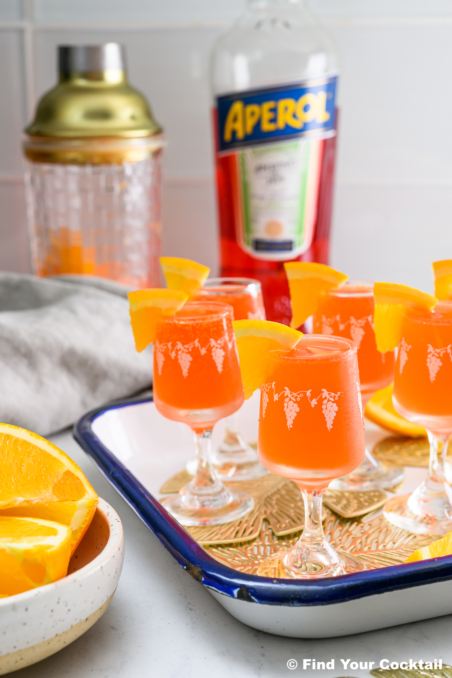 A tray of orange Aperol cocktails in small glasses with orange slices, a shaker, a bottle of Aperol, and a bowl of orange wedges in the background.