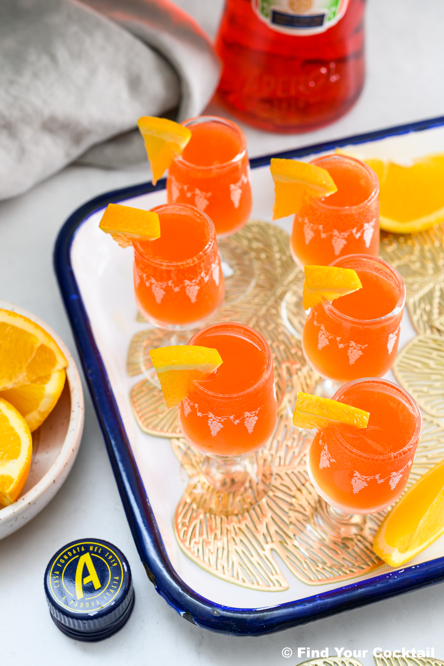 Six orange-colored shots with orange wedges on a tray, a bottle of Aperol, and a bowl of orange slices visible nearby.
