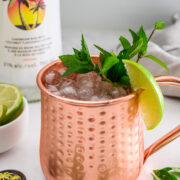 A copper mug filled with crushed ice, garnished with a lime wedge and mint, sits near a bottle of Malibu rum and a small bowl of lime slices.