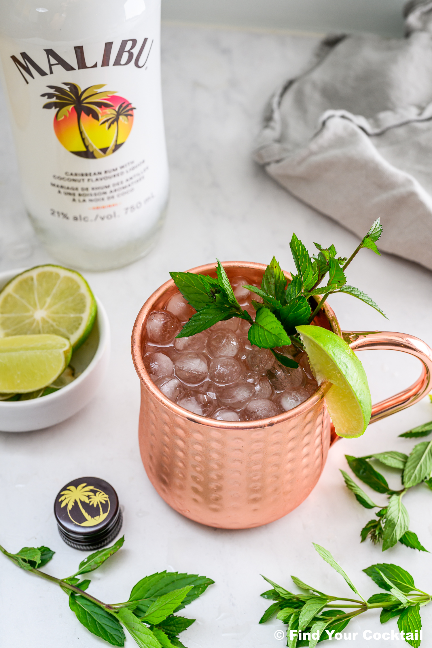 A copper mug filled with ice, garnished with mint and a lime wedge, sits next to a Malibu bottle, a bowl of lime slices, and mint sprigs on a marble surface.