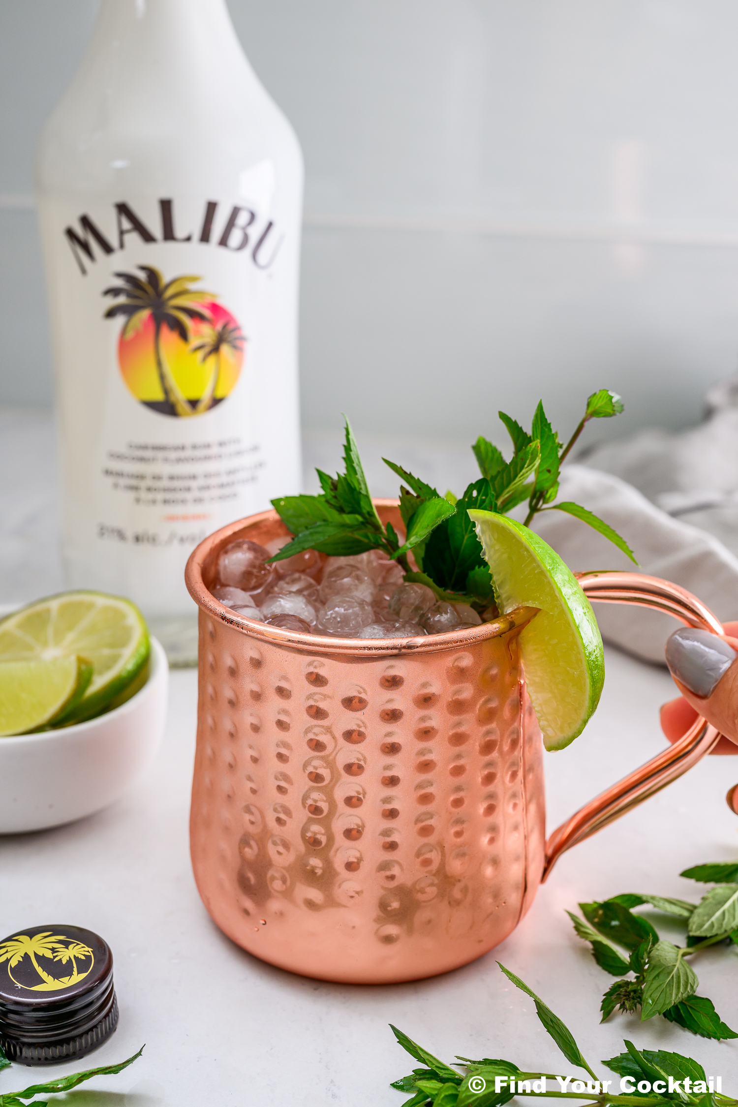 A copper mug filled with ice, garnished with mint and lime, sits in front of a Malibu rum bottle, with lime wedges and mint leaves nearby.