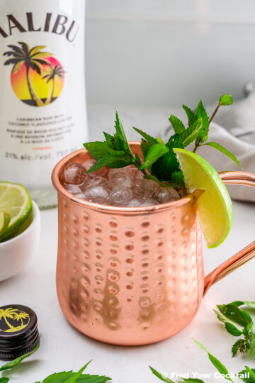A copper mug filled with crushed ice, garnished with a lime wedge and mint, sits near a bottle of Malibu rum and a small bowl of lime slices.