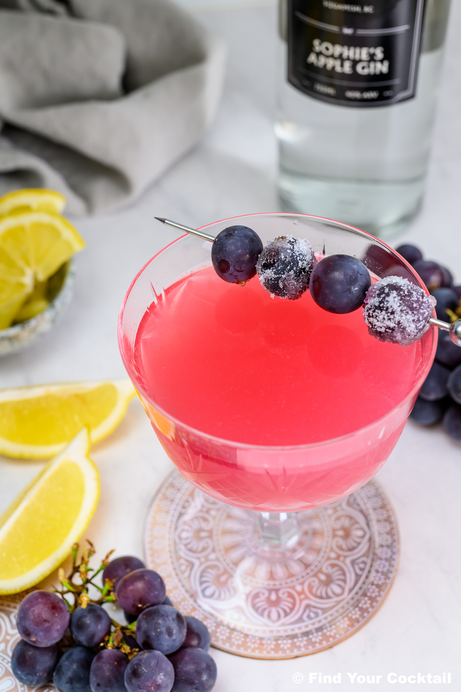 A pink cocktail in a glass garnished with sugared grapes on a skewer, surrounded by fresh grapes, lemon wedges, and a bottle of gin in the background.