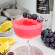 A pink cocktail in a glass, garnished with grapes on a skewer, sits beside a gin bottle, lemon slices, and loose grapes on a white surface.
