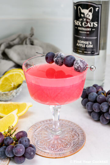 A pink cocktail in a glass, garnished with grapes on a skewer, sits beside a gin bottle, lemon slices, and loose grapes on a white surface.