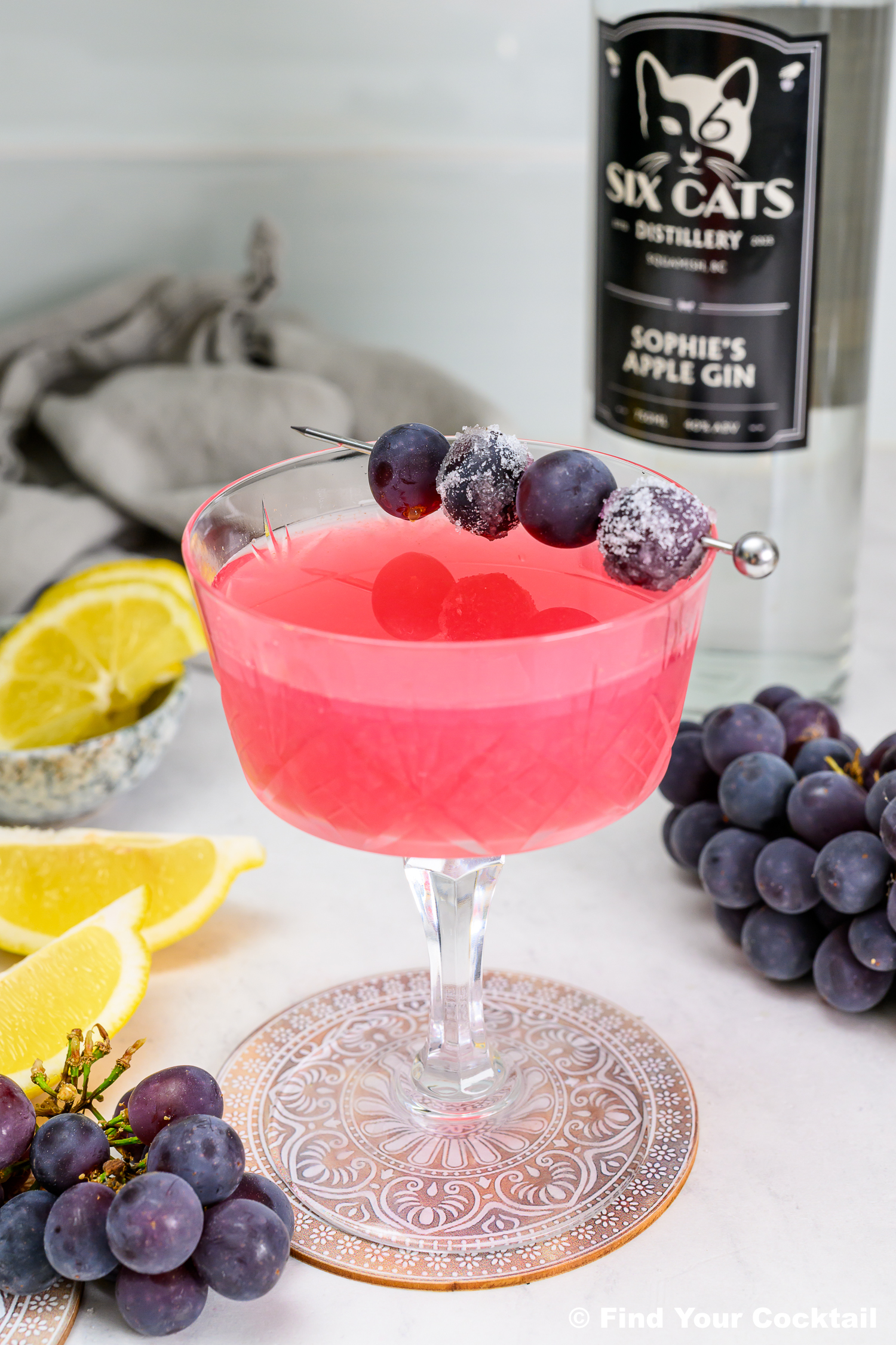 A pink cocktail in a glass, garnished with grapes on a skewer, sits beside a gin bottle, lemon slices, and loose grapes on a white surface.