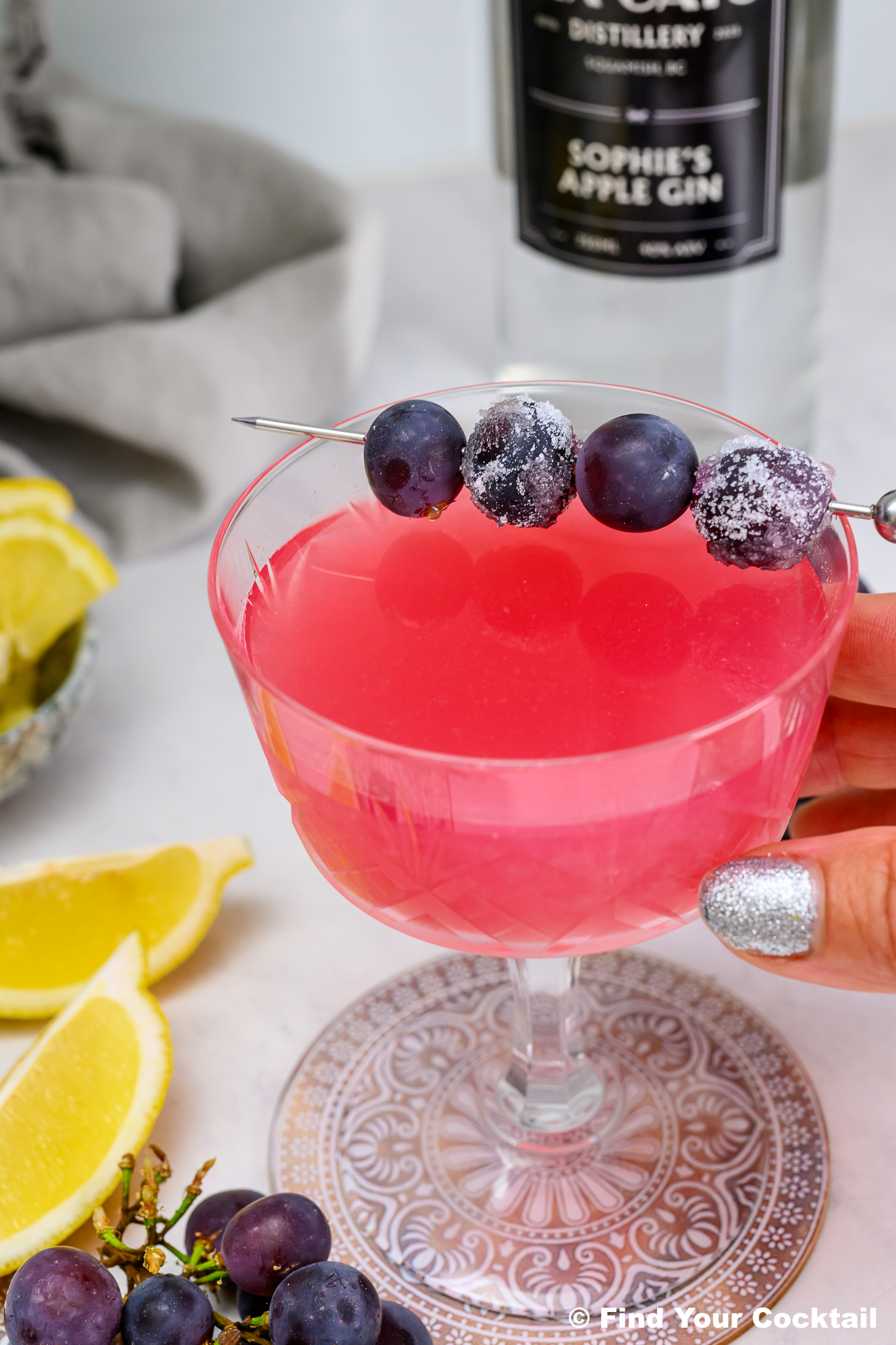 A hand holds a pink cocktail in a coupe glass, garnished with three grapes on a skewer. Lemon wedges and grapes are nearby, with a bottle labeled apple gin in the background.