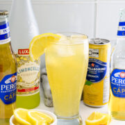 A tall glass of lemon cocktail with ice and a lemon slice, surrounded by bottles of Peroni beer, Limoncello, San Pellegrino Limonata, and a bowl of lemon wedges on a white counter.