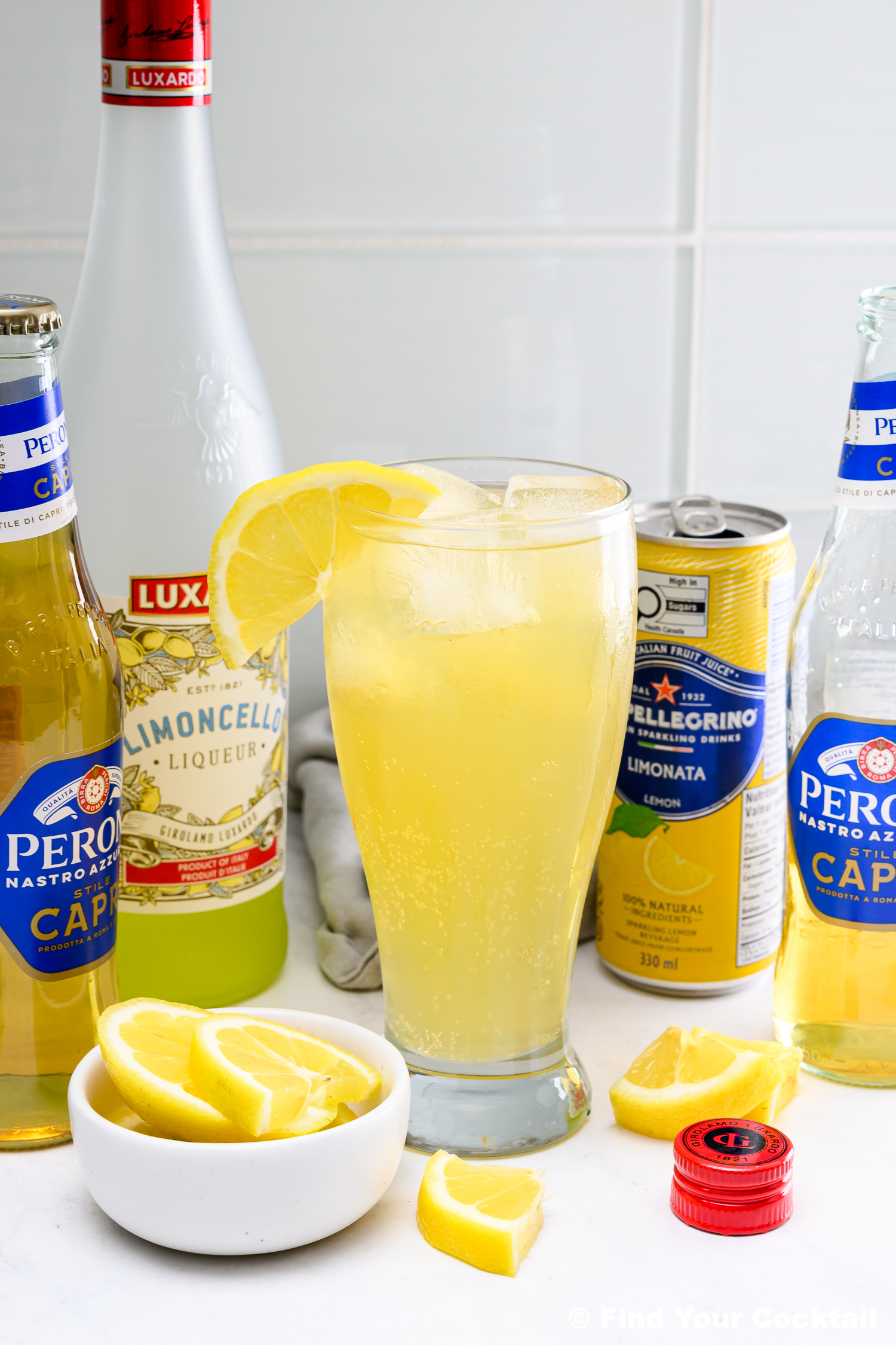A tall glass of lemon cocktail with ice and a lemon slice, surrounded by bottles of Peroni beer, Limoncello, San Pellegrino Limonata, and a bowl of lemon wedges on a white counter.