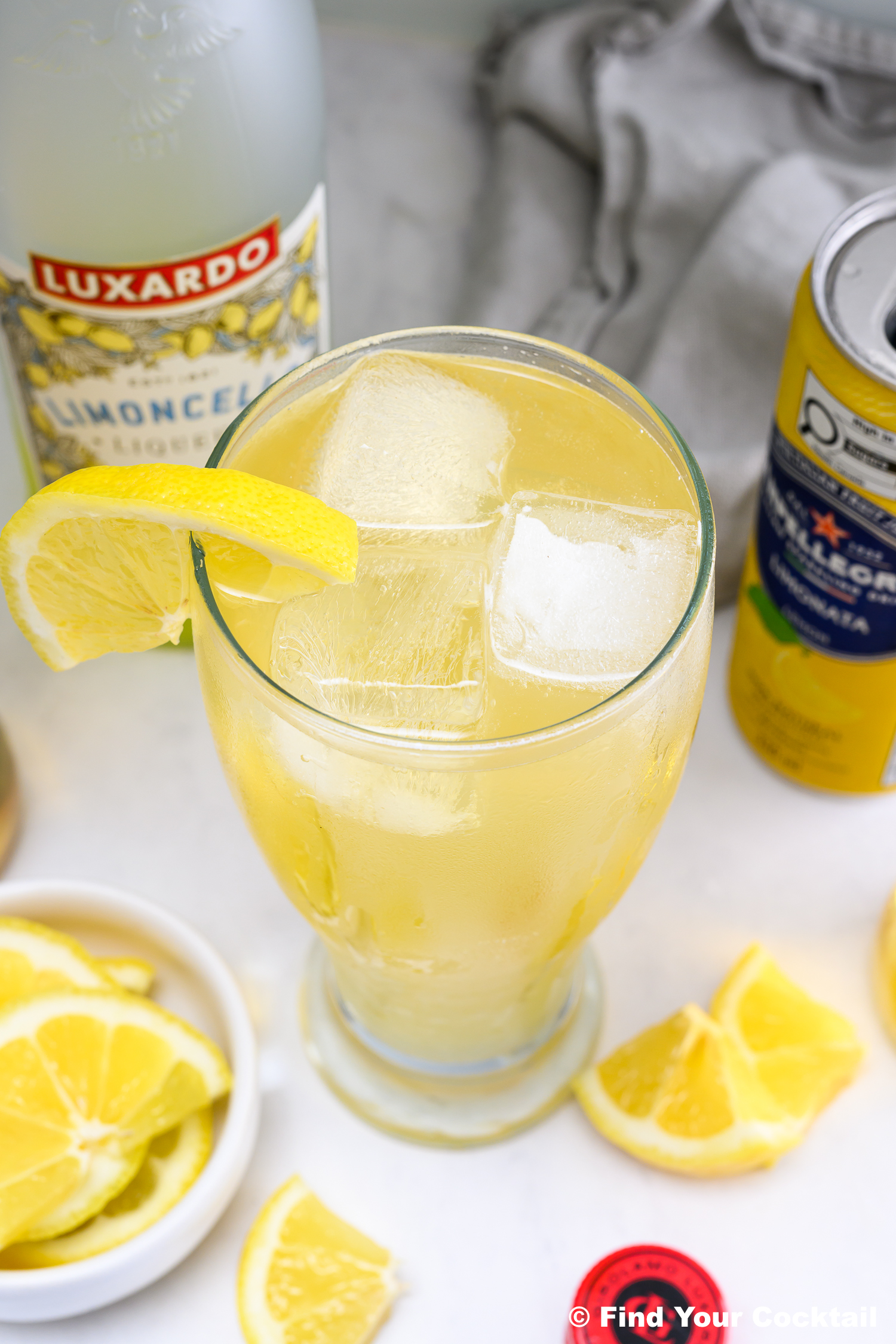 Tall glass of yellow cocktail with ice and lemon wedge, surrounded by sliced lemons, a can, and a bottle labeled Luxardo Limoncello..