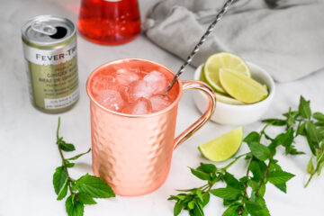 A copper mug filled with ice and a metal straw, next to ginger beer, lime wedges in a bowl, and mint sprigs on a white surface.