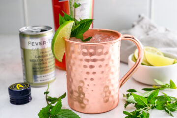 A copper mug filled with ice, garnished with a lime wedge and mint sprig, sits beside a can of ginger beer, a bottle cap, and fresh mint leaves.