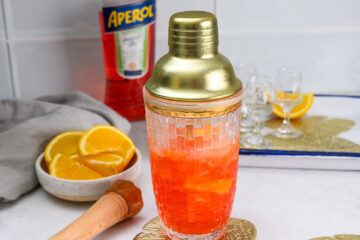 A cocktail shaker filled with orange-red liquid sits on a counter near sliced oranges, a muddler, shot glasses, and a bottle of Aperol.