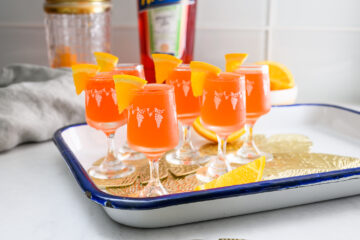 Six small orange cocktails with orange slices garnish are arranged on a white tray with a blue rim, with a bottle, jar, and orange in the background.