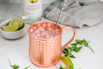 A copper mug filled with crushed ice and a metal straw sits on a counter with lime wedges, mint leaves, and a gray cloth nearby.